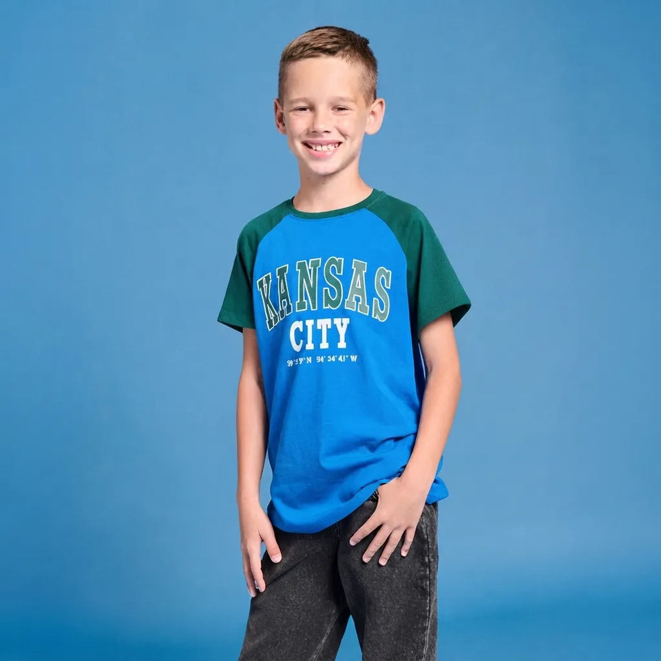 A smiling young boy wearing a blue and green raglan t-shirt with 'KANSAS CITY BIRTHDAY' printed on the front, standing against a solid blue studio background.