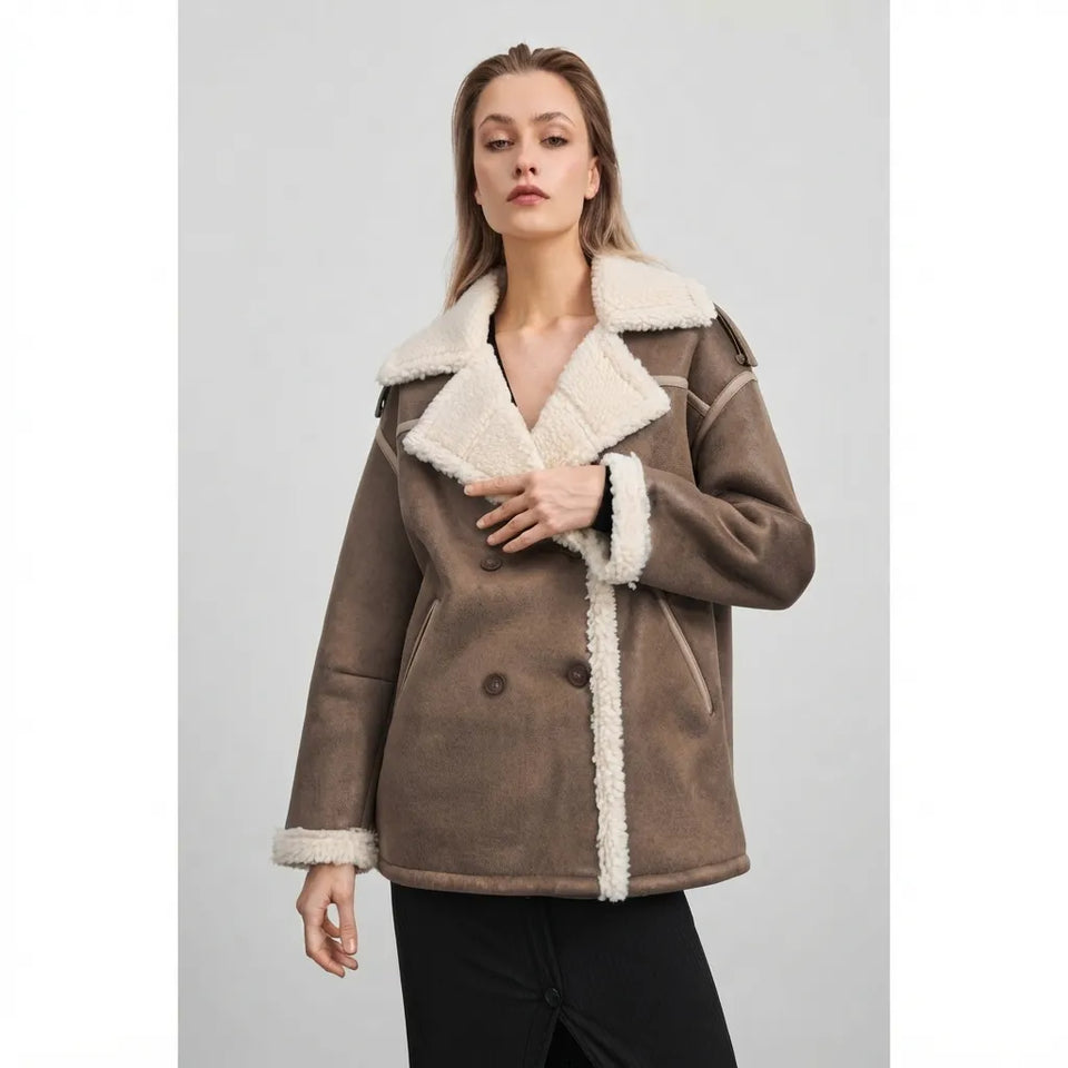 A woman modeling a brown shearling-lined jacket with a cream-colored collar, trim, and cuffs, paired with black pants, against a solid light gray background.