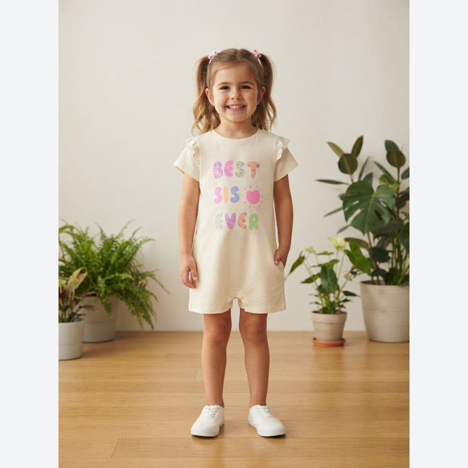 A young girl with blonde pigtails stands smiling in a cream-colored short-sleeve romper with 'BEST BFF EVER' printed in pastel pink, green, and blue letters. She is wearing white sneakers in a brightly lit room with a wooden floor and potted plants in the background.