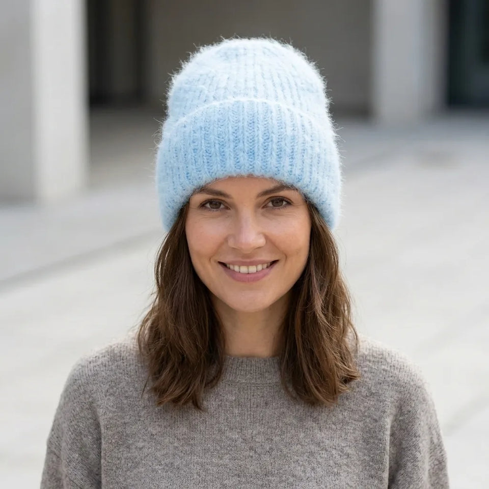 A smiling woman wearing a light blue fuzzy knit beanie hat and a gray knit sweater, photographed outdoors against a softly blurred neutral background.