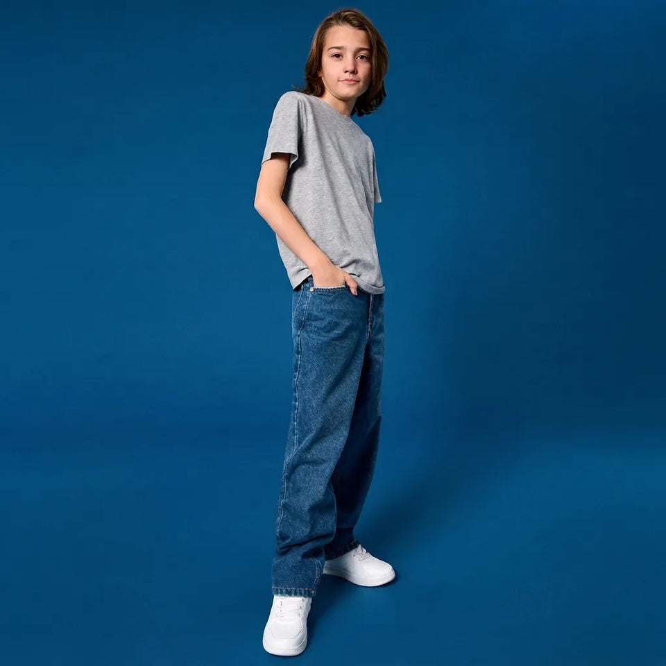 A child model wearing a heather gray short-sleeve t-shirt, loose-fit blue jeans, and white sneakers, standing against a solid blue studio background.