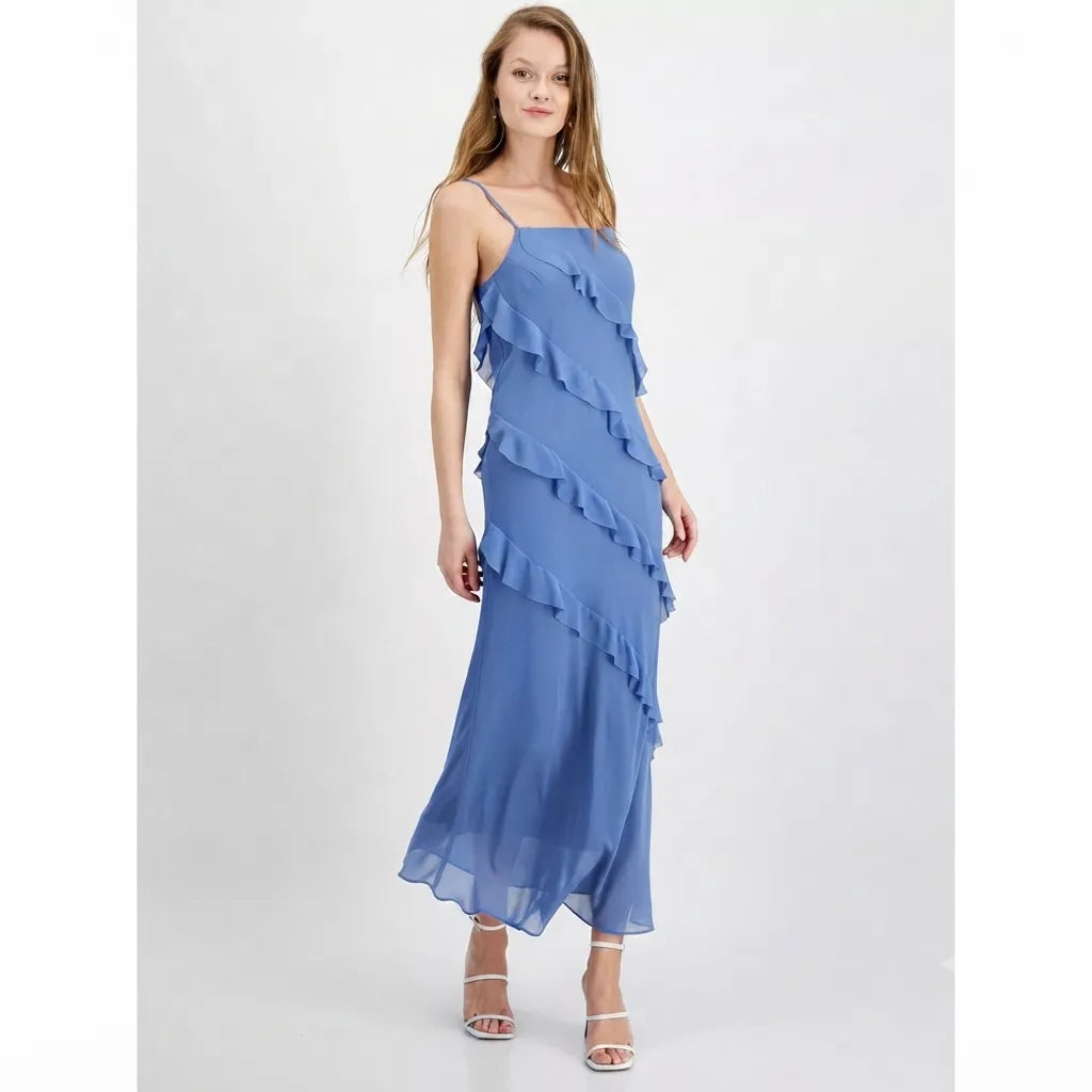 A woman modeling a blue ruffled midi dress with spaghetti straps and a flowing chiffon-like skirt, paired with white strappy high-heeled sandals, against a light gray studio background.