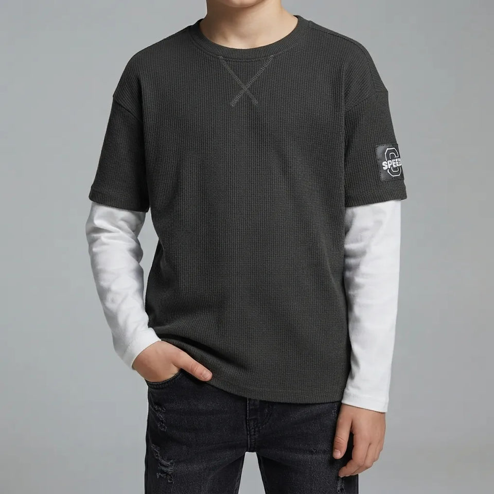 A child wearing a dark gray waffle-knit long-sleeve shirt with a white layered long-sleeve shirt underneath, paired with dark jeans, against a solid gray studio background.