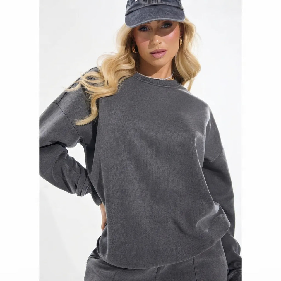 A woman wearing a dark gray crewneck sweatshirt and matching sweatpants, paired with a dark gray baseball cap, photographed against a plain white studio background.