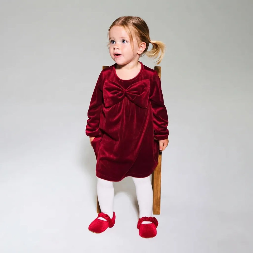 A toddler girl wearing a long-sleeve burgundy velvet dress with a large bow at the chest, white tights, and red shoes, standing on a wooden stool against a light gray studio background.