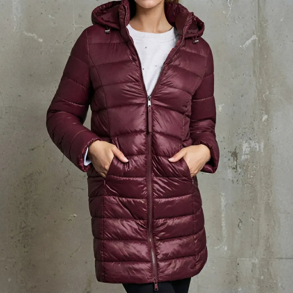 A woman wearing a shiny, quilted hooded puffer jacket in a deep burgundy/maroon color, standing against a textured gray concrete wall.