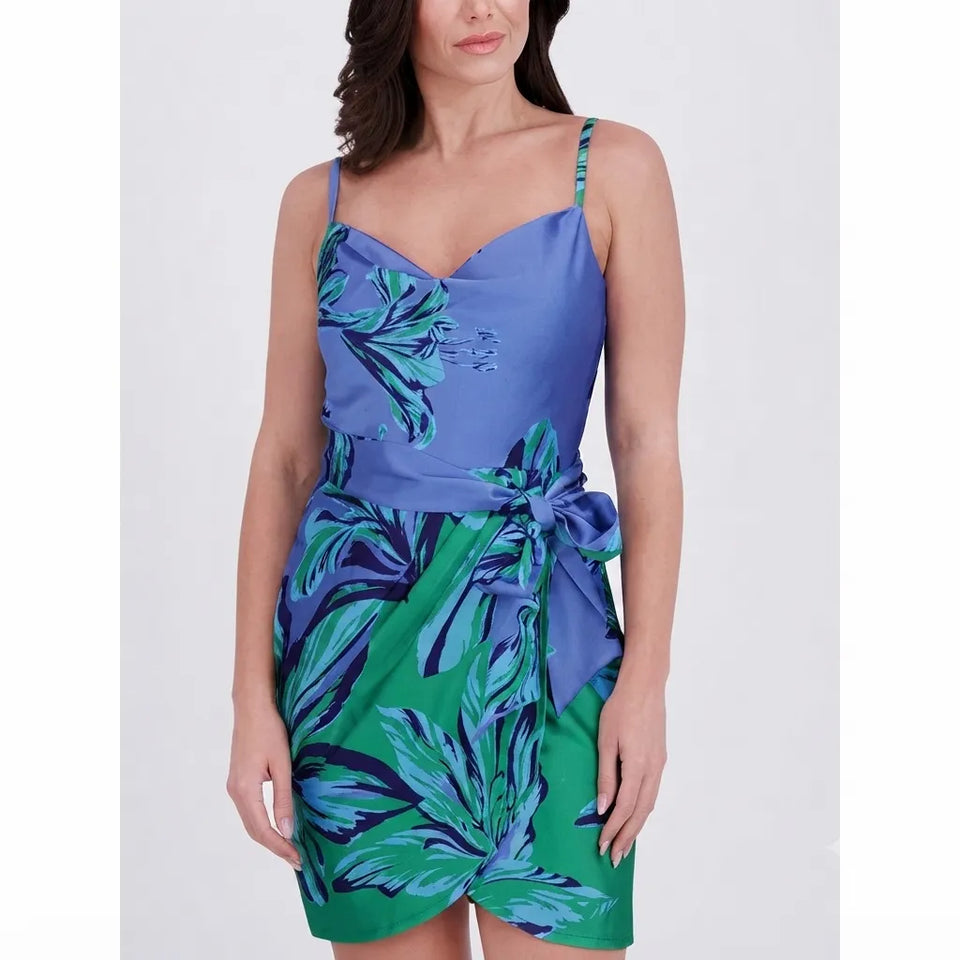 A woman wearing a sleeveless wrap-style mini dress with thin straps, a blue bodice, and a green tropical leaf print, photographed against a light gray studio background.