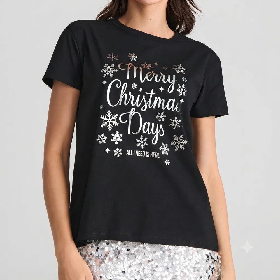 A woman wearing a black short-sleeve graphic T-shirt with 'Merry Christmas Days' and 'ALL I NEED IS HERE' printed in white and metallic script, surrounded by white snowflakes. The model is also wearing silver sequined bottoms against a light gray studio background.