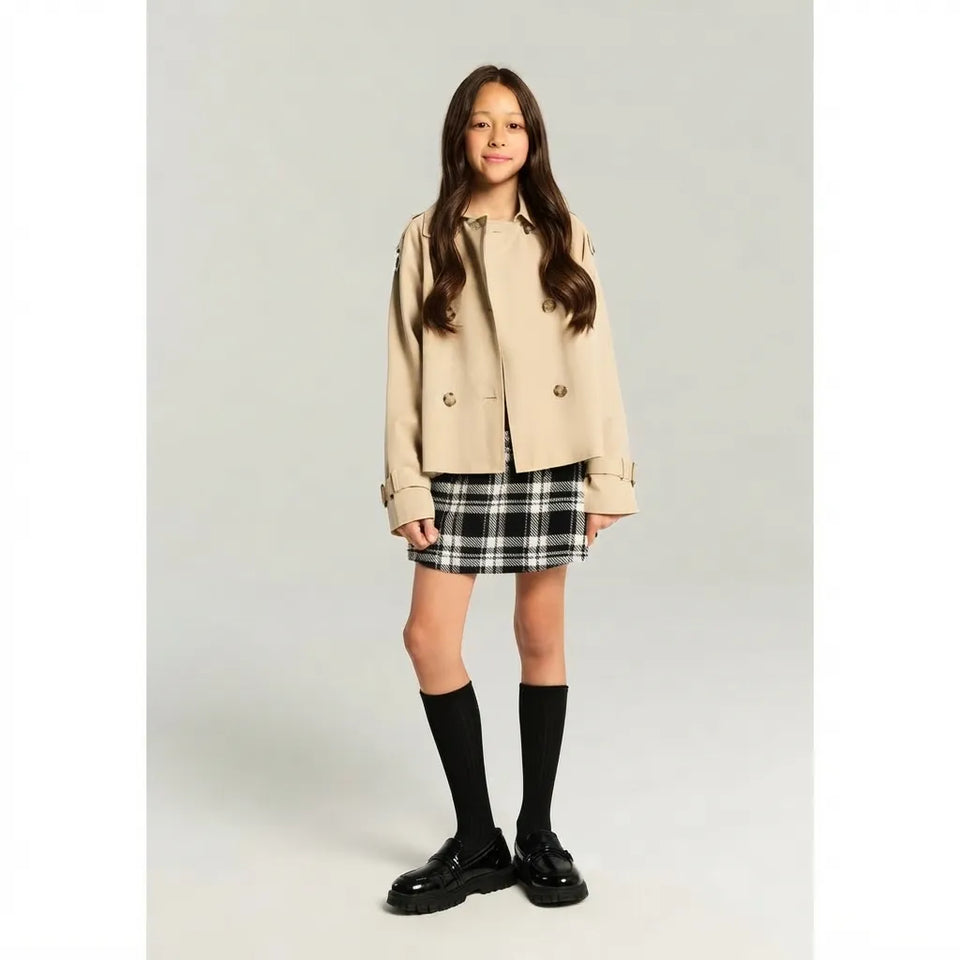 A young girl stands against a light gray studio background wearing a beige double-breasted jacket, a black and white plaid mini skirt, black knee-high socks, and black leather shoes.