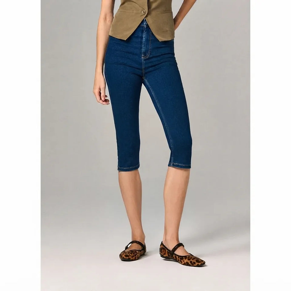 A model wearing dark blue cropped capri jeans, paired with a tan top and leopard-print flat shoes, standing against a light gray studio background.