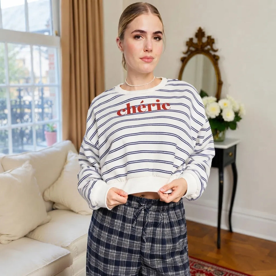 A woman wearing a cropped white sweatshirt with thin navy horizontal stripes and the word 'chéric' in red across the chest, paired with navy plaid pants, standing in a bright living room.