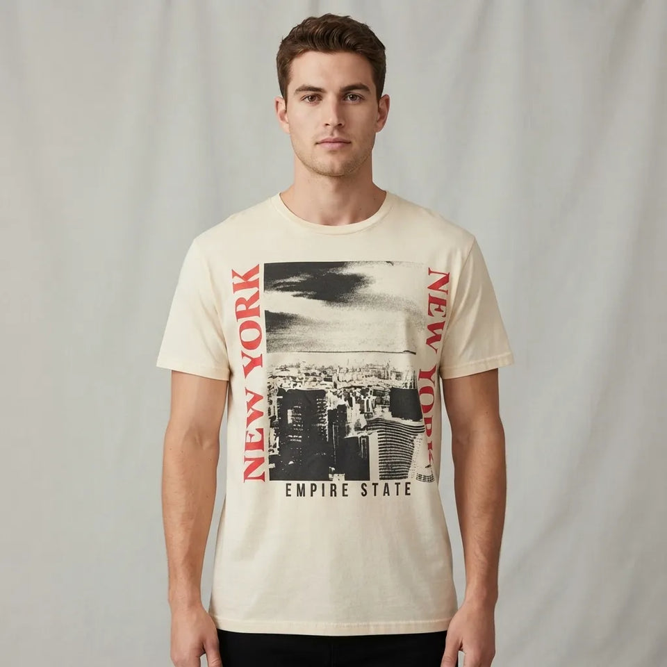 A man wearing a cream-colored graphic t-shirt with a black and white cityscape print and red vertical 'NEW YORK' text, set against a neutral gray backdrop.