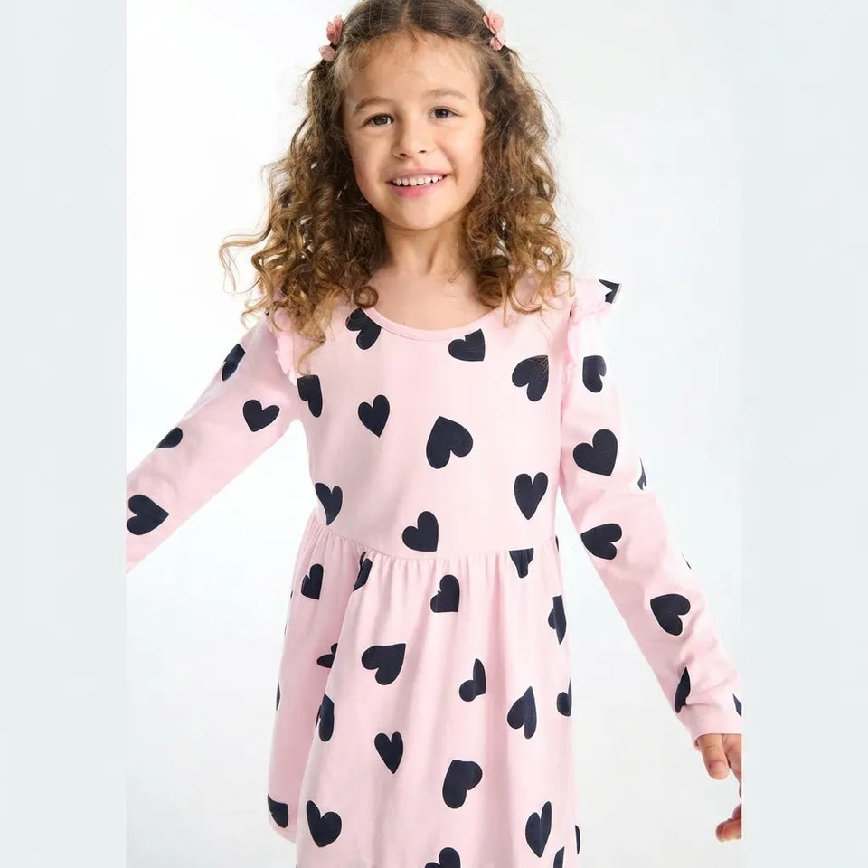 A young girl with curly brown hair wearing a light pink long-sleeve dress with a black heart pattern, photographed against a plain white studio background.