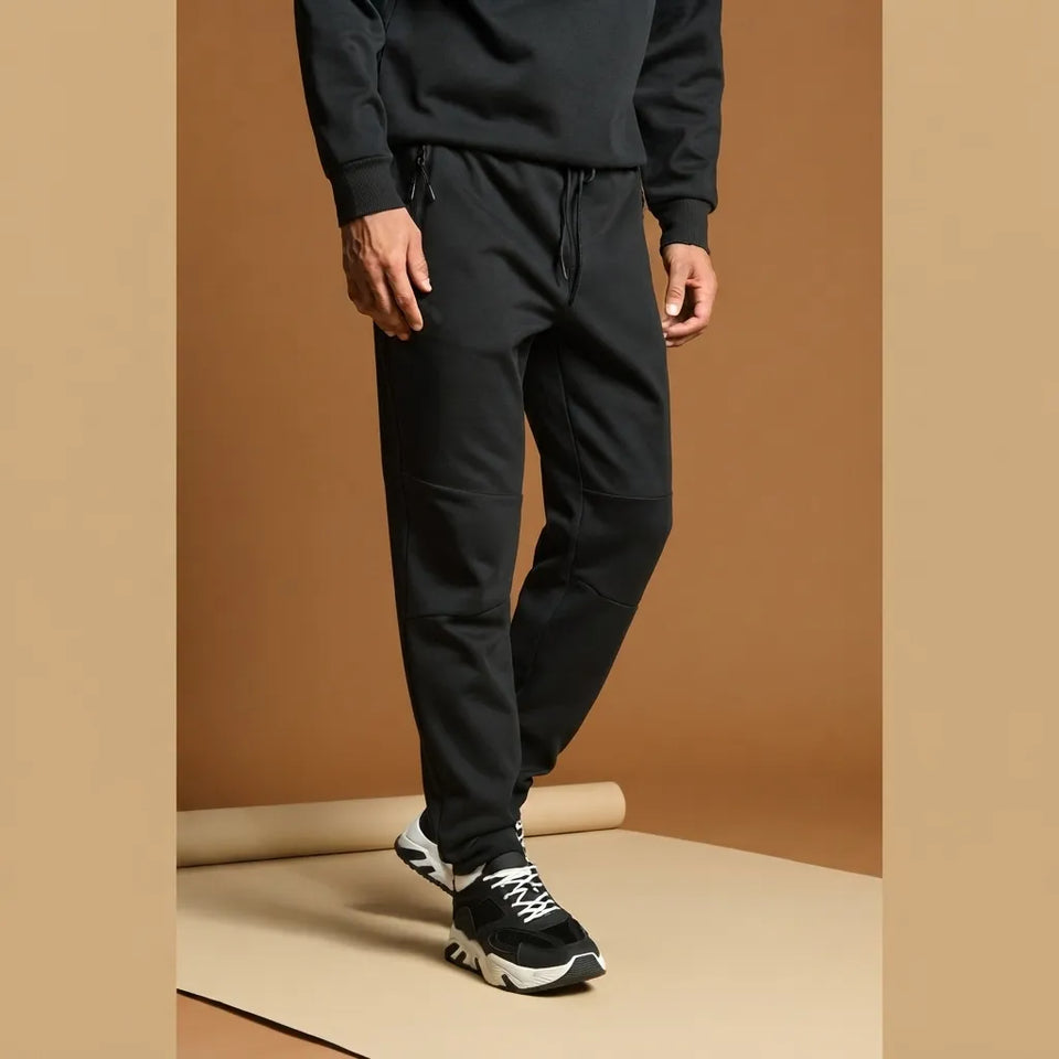 A person wearing black jogger-style sweatpants with a drawstring waist, paired with a black sweatshirt and black-and-white sneakers, standing against a warm tan studio backdrop.