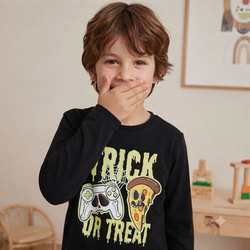 A young boy with brown hair wearing a black long-sleeve shirt with a 'Trick or Treat' graphic featuring a video game controller and a pizza slice, standing in a softly lit indoor room.
