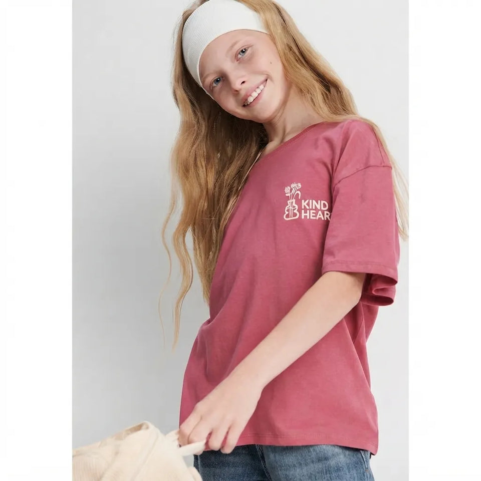 A young girl with long blonde hair wearing a dusty rose pink t-shirt with a white 'KIND HEART' graphic on the left chest, paired with a white headband and blue jeans, holding a beige bag against a light gray background.