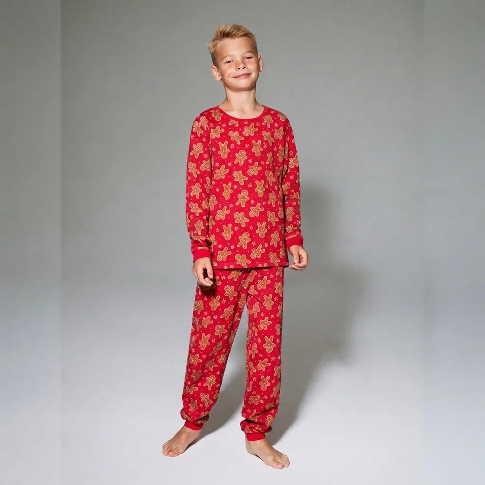 A young boy stands barefoot against a light gray studio background, wearing a red long-sleeve pajama set with a repeating gold floral pattern.