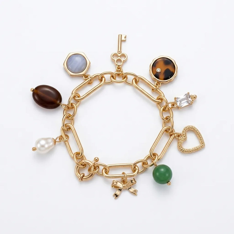 A gold paperclip chain charm bracelet on a white background, featuring multiple dangling charms including a key, a heart, a green bead, a pearl, a brown oval stone, a tortoiseshell oval, a blue-gray hexagon, and a small silver crystal charm.