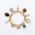 A gold paperclip chain charm bracelet on a white background, featuring multiple dangling charms including a key, a heart, a green bead, a pearl, a brown oval stone, a tortoiseshell oval, a blue-gray hexagon, and a small silver crystal charm.