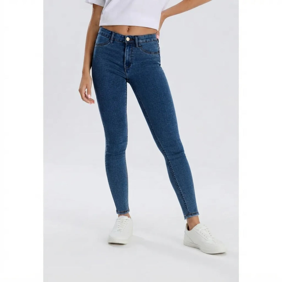 A model wearing a pair of high-waisted, skinny blue jeans with a white crop top and white sneakers, shown from the waist down against a light gray studio background.