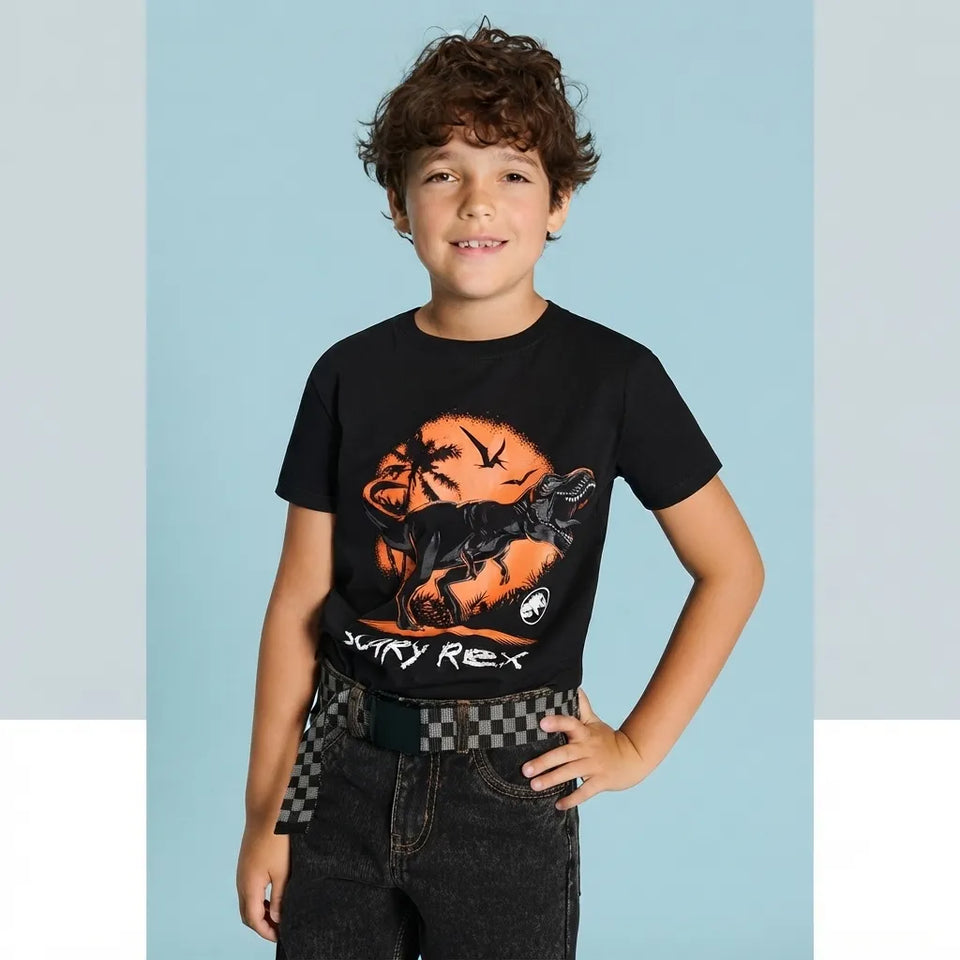 A young boy with curly brown hair wearing a black graphic t-shirt with an orange and black 'Scary Rex' dinosaur design, paired with dark jeans and a checkered belt, posing against a light blue background.