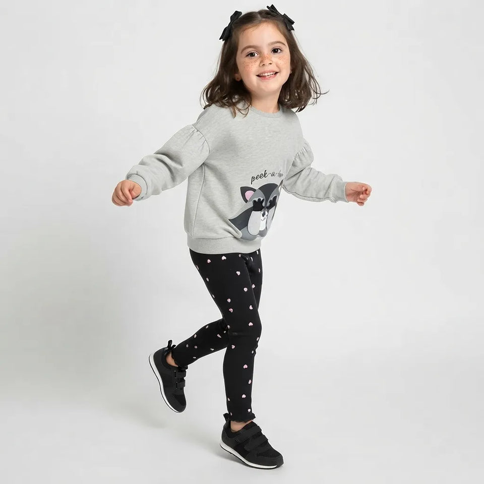 A young girl smiling and posing in a light gray sweatshirt with a dog graphic and the text 'pet a dog', paired with black leggings featuring small pink hearts and black sneakers, set against a plain light gray studio background.