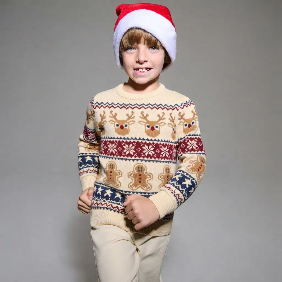 A young boy wearing a festive Christmas sweater and Santa hat, standing against a neutral gray studio background.