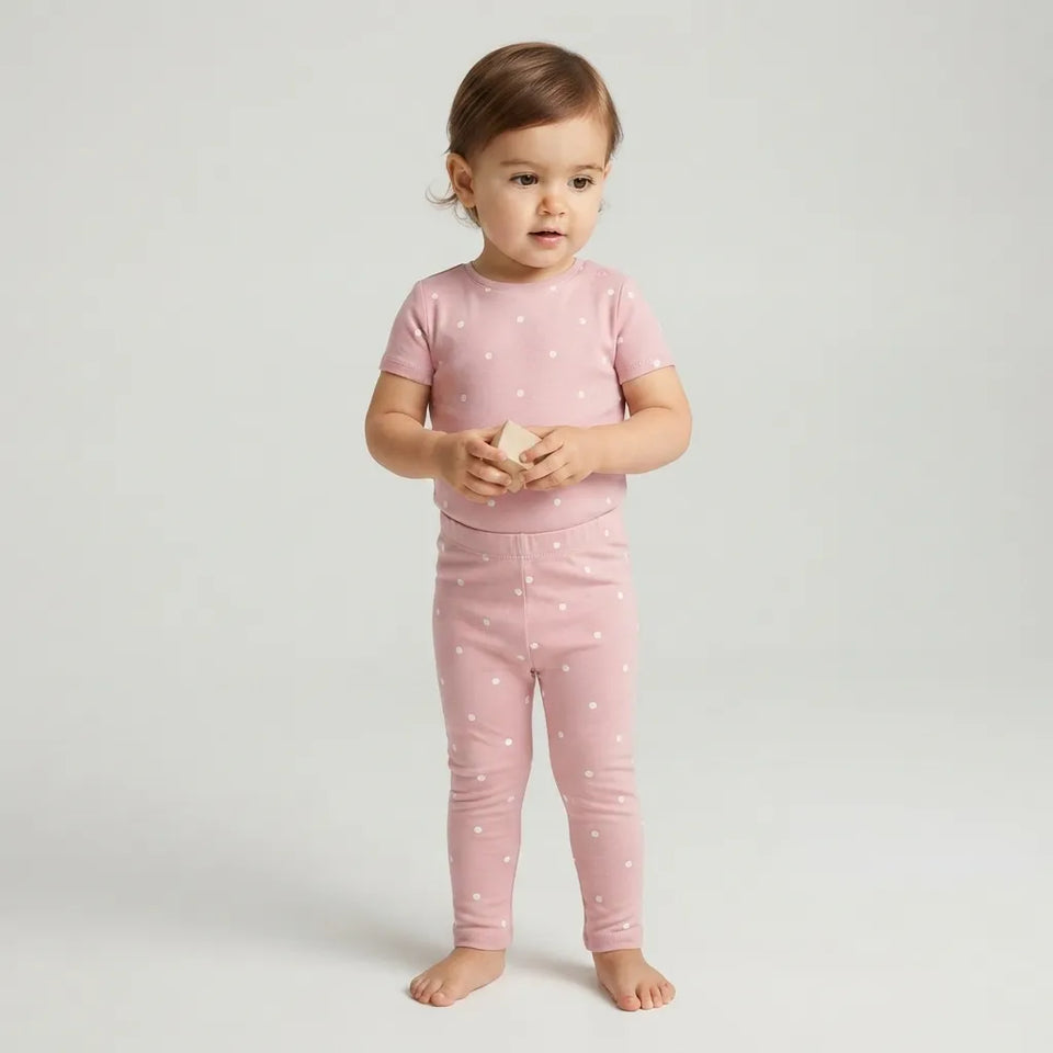 A toddler standing barefoot against a light gray studio background, wearing a matching pink short-sleeve top and leggings set with small white polka dots.