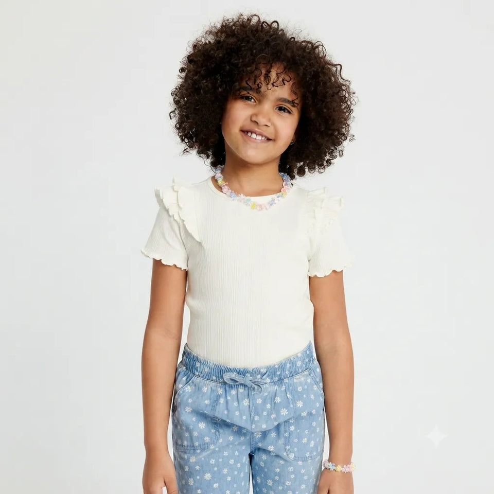A young girl with curly brown hair wearing a cream ribbed short-sleeve top with ruffle trim and light blue polka dot pants, standing against a plain white background.