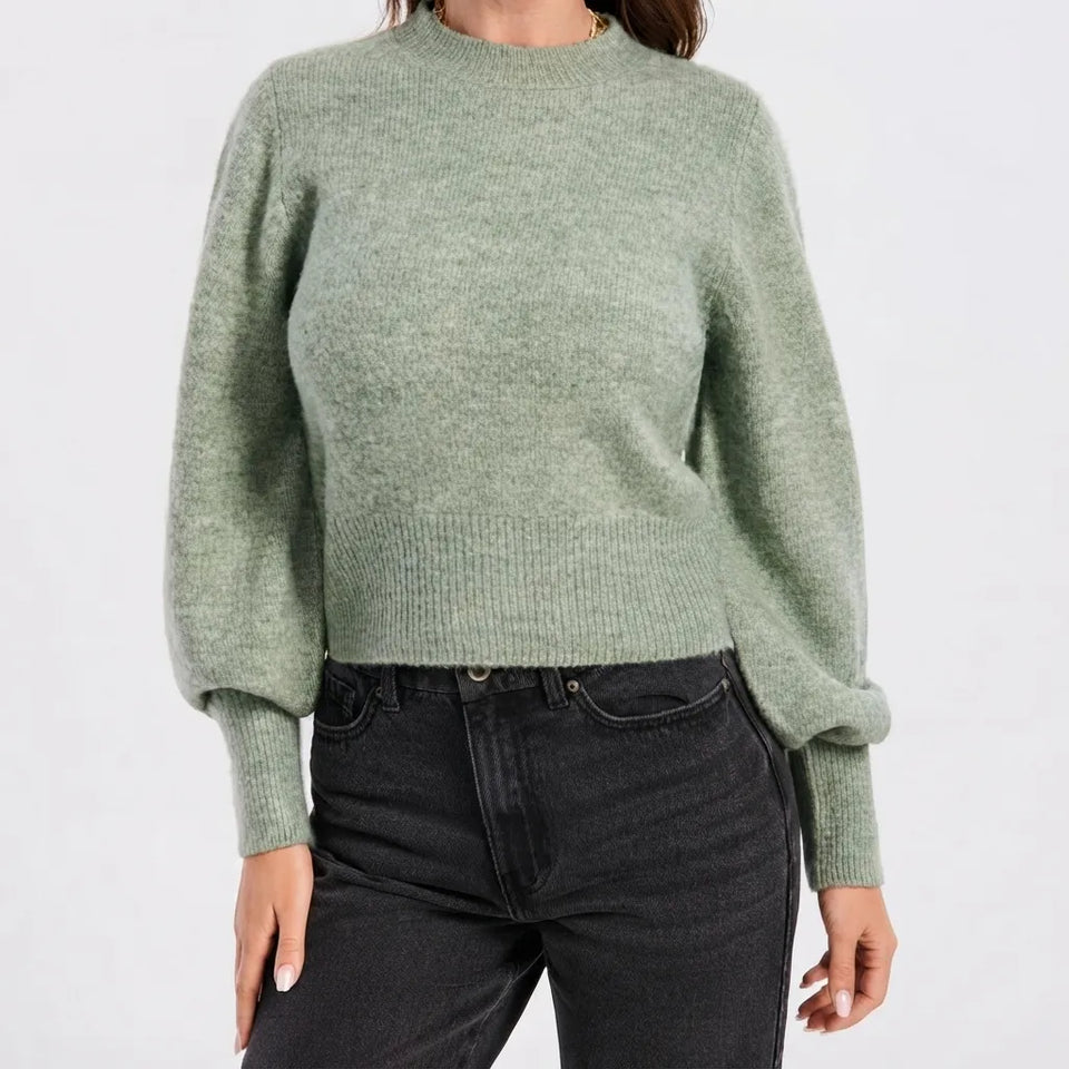 A woman wearing a light sage green knit sweater with balloon sleeves and dark gray jeans, shown from the neck down against a plain white background.