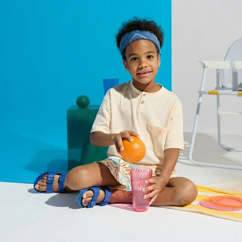 A young child sits cross-legged on a white floor, holding an orange and a pink cup. The child wears a cream short-sleeve top, patterned shorts, blue sandals, and a blue patterned headband.