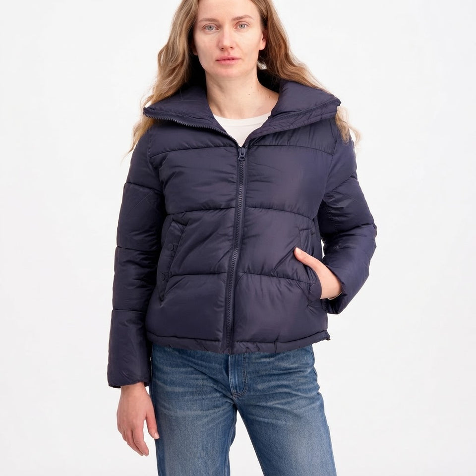 A woman wearing a dark navy blue puffer jacket with a high collar, front zipper, and horizontal quilting, paired with blue jeans, against a plain white background.