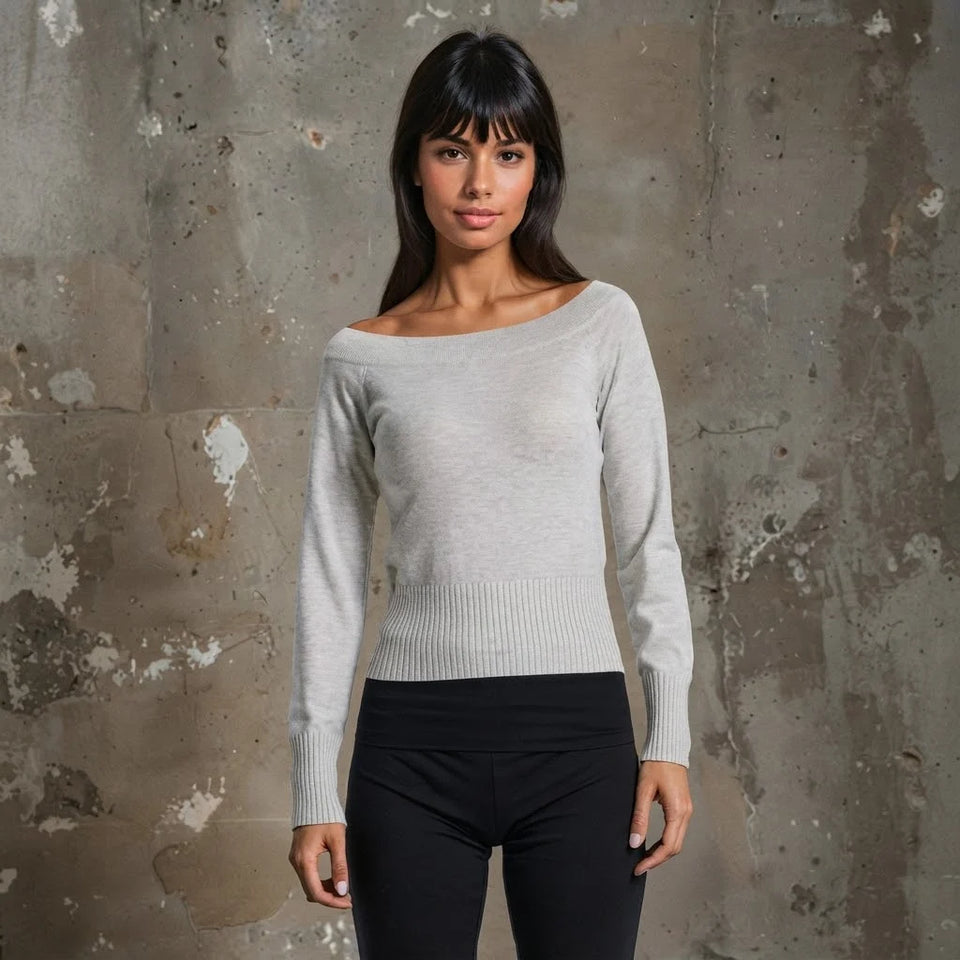 A woman modeling a light gray off-the-shoulder long-sleeve knit sweater with black pants, standing against a textured concrete wall.