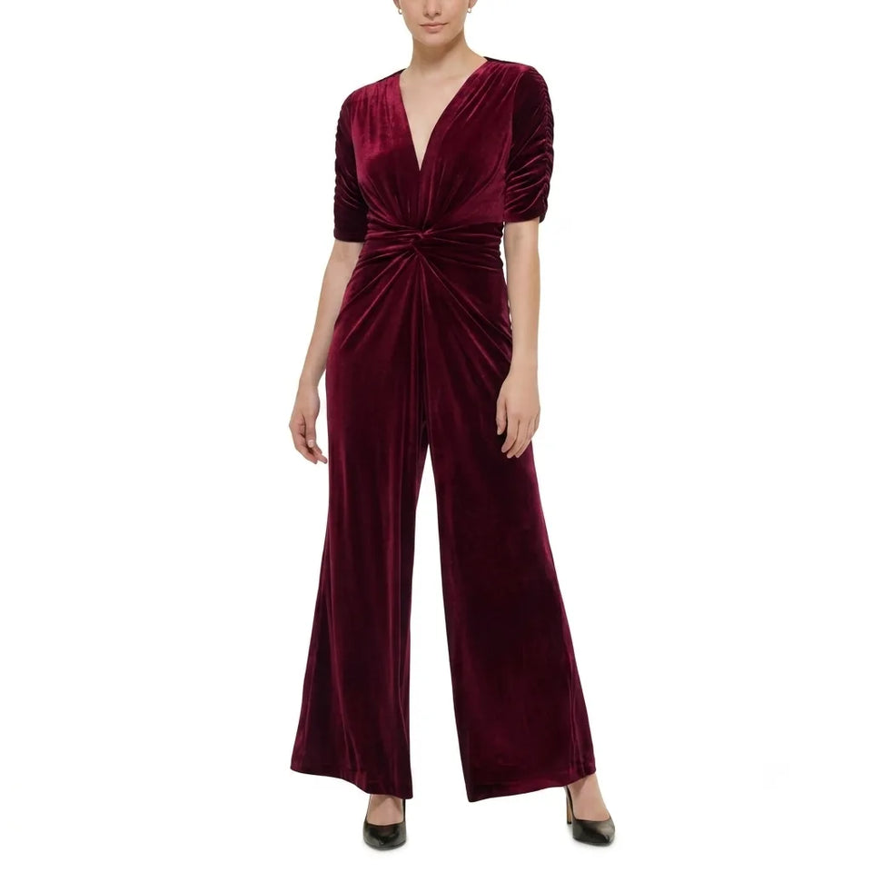 A woman modeling a deep burgundy velvet jumpsuit with short sleeves, a wrap-style V-neckline, and a twisted waist detail, paired with black pointed shoes against a plain white background.