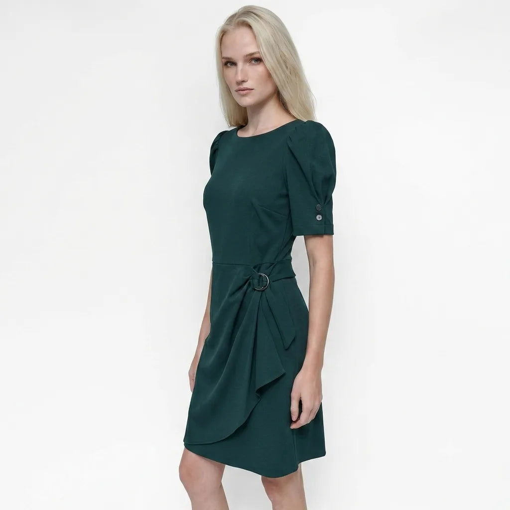 A blonde model wearing a dark green short-sleeve dress with puff sleeves, a matching tie belt, and a wrap-style skirt, photographed against a plain white studio background.