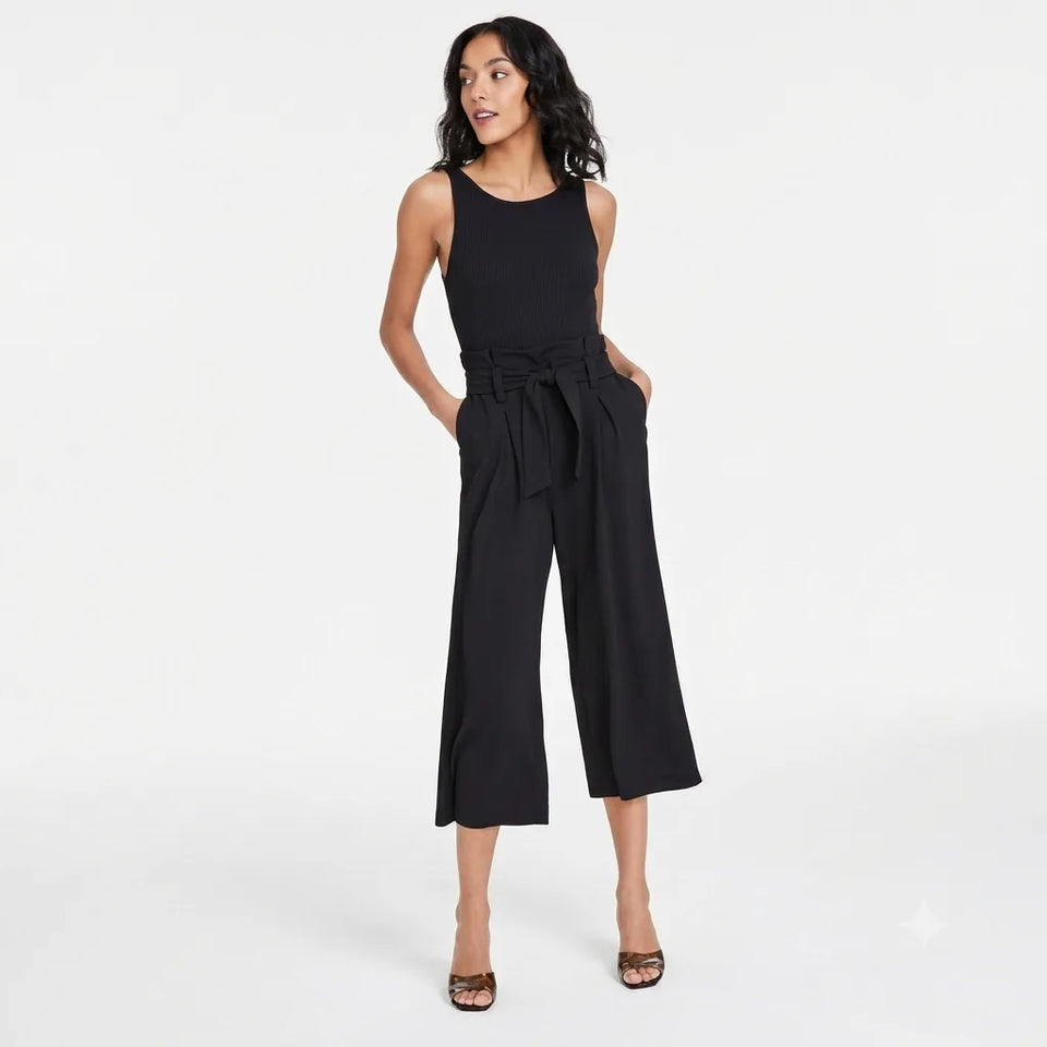 A woman modeling a black sleeveless wide-leg cropped jumpsuit with a matching waist tie, paired with brown heeled sandals, against a plain white studio background.