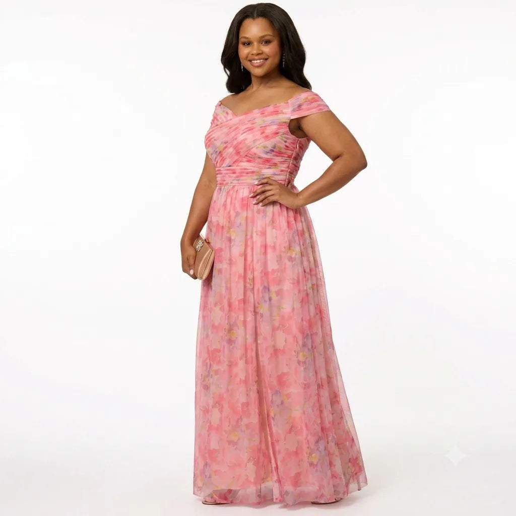 A woman modeling a pink off-the-shoulder maxi dress with a floral print and a matching self-tie sash at the waist, holding a small beige clutch against a plain white studio background.