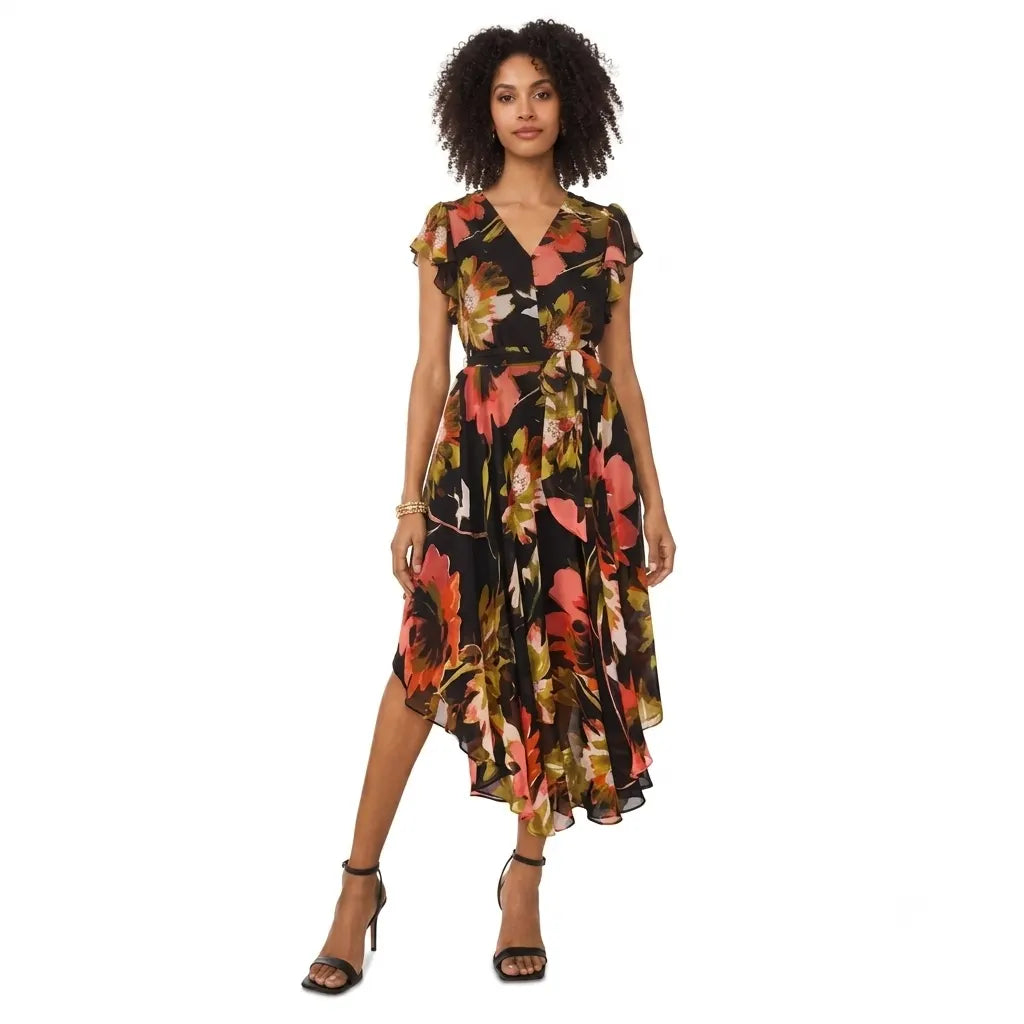 A woman modeling a short-sleeve midi dress with a black base and a vibrant floral print in orange, pink, and yellow, paired with black strappy high-heeled sandals against a plain white background.