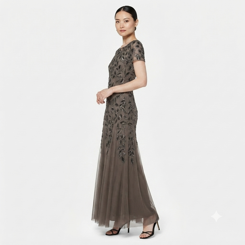 A woman modeling a floor-length evening gown in a dark taupe-gray color with intricate floral embroidery and sheer tulle detailing, set against a plain light gray background.