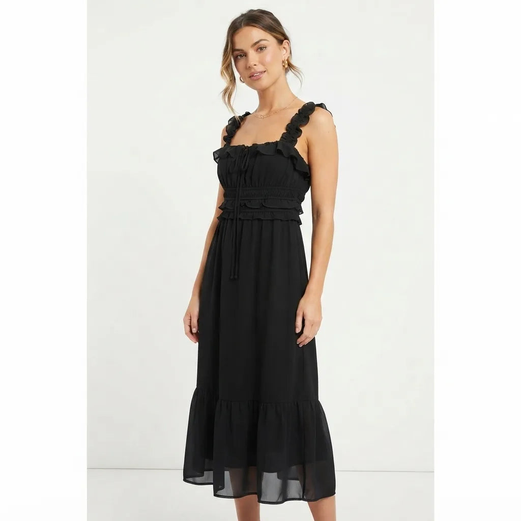 A woman modeling a black midi dress with ruffled shoulder straps, a gathered sweetheart neckline, a matching ruffled waistband, and a tiered skirt against a plain white studio background.