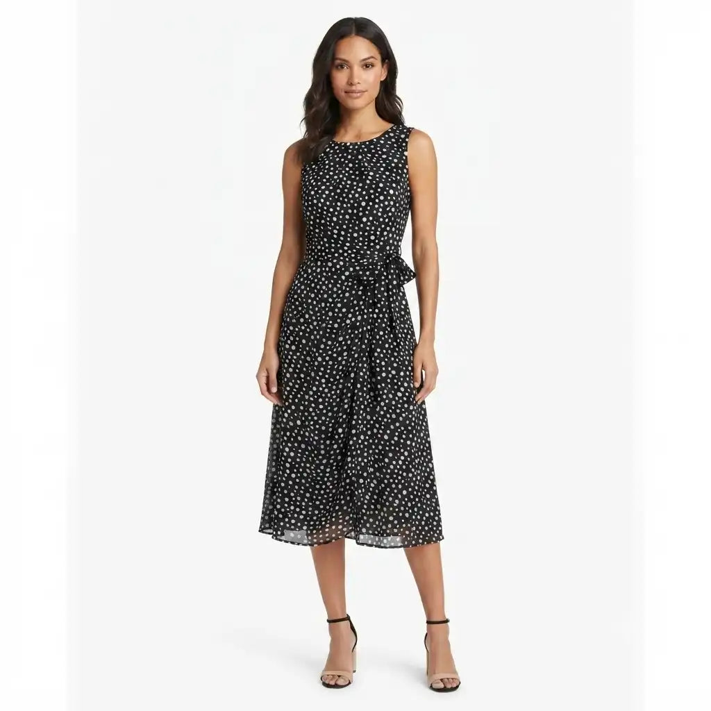 A woman modeling a sleeveless black midi dress with a white polka dot pattern and a matching fabric tie belt at the waist, set against a plain light gray studio background.