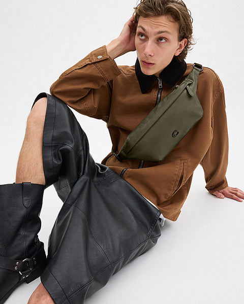 Man wearing a brown jacket and black pants with a green bag, sitting on a white background
