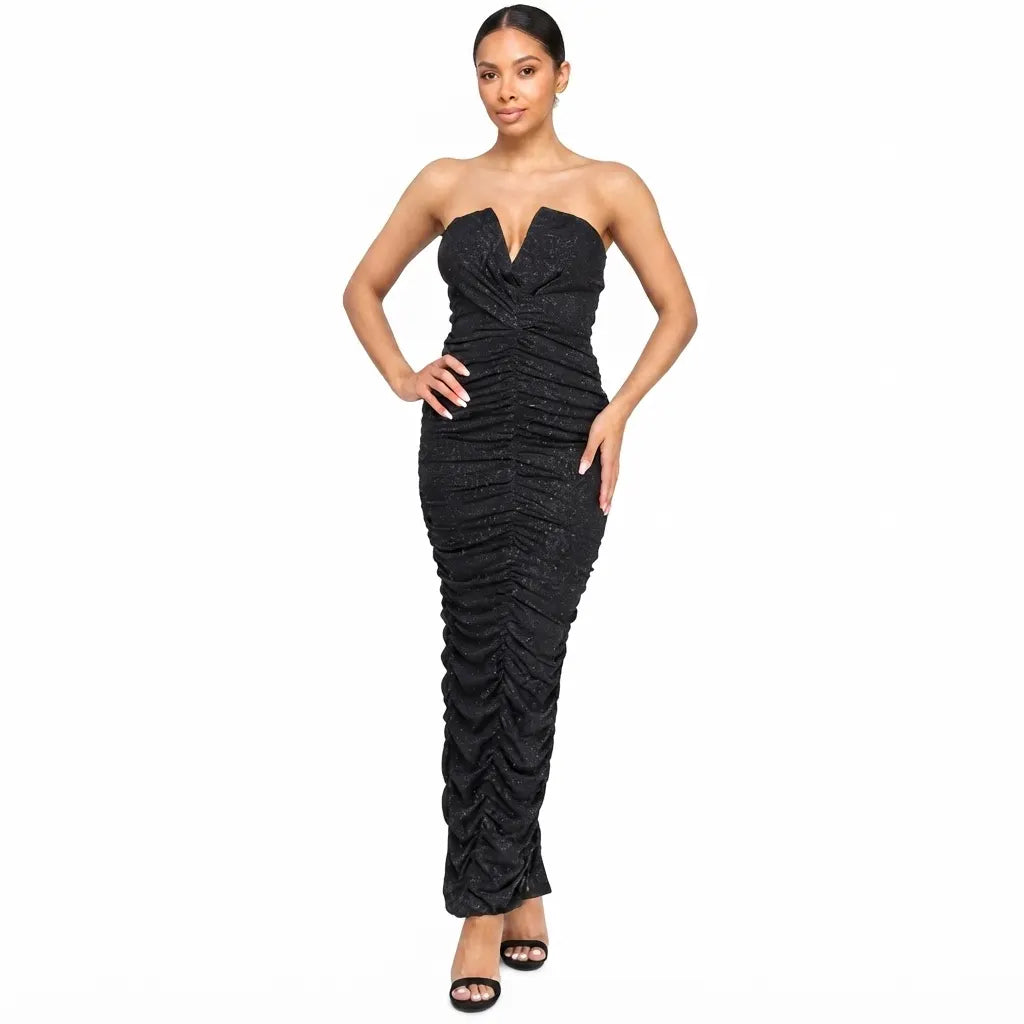 A woman modeling a strapless black ruched maxi dress with a deep V-neckline and fitted silhouette, paired with black heeled sandals, against a plain white background.