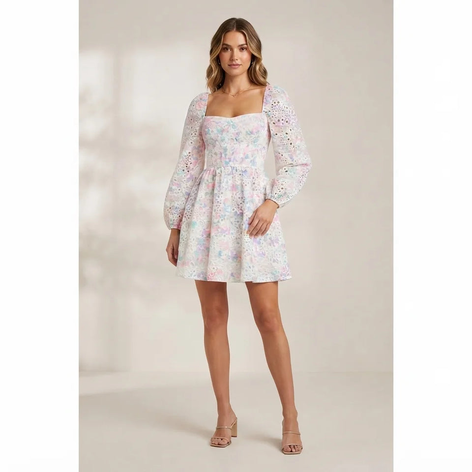 A woman modeling a short white puff-sleeve dress with a pastel multicolor floral print, photographed against a warm beige studio background.
