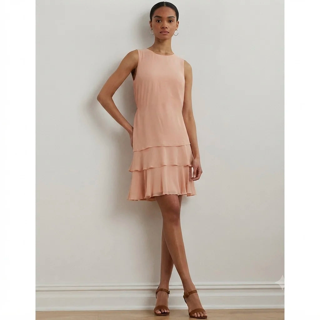 A woman models a sleeveless, tiered mini dress in a soft peach-pink color, standing against a plain off-white wall.