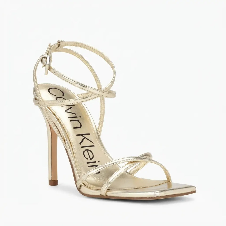A single metallic gold Calvin Klein high-heeled sandal with thin wrap-around ankle straps and a stiletto heel, shown on a clean white background.