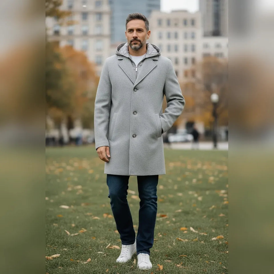 A man wearing a light gray mid-length overcoat with a gray-and-white patterned hood, dark blue jeans, and white sneakers, standing on a grassy park lawn with blurred buildings in the background.