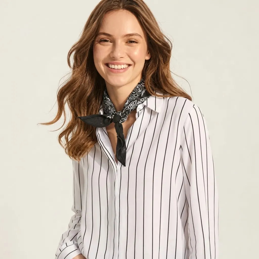 A woman with wavy brown hair smiling, wearing a white long-sleeve button-up shirt with thin black vertical pinstripes and a black-and-white patterned neck scarf, set against a plain light beige studio background.