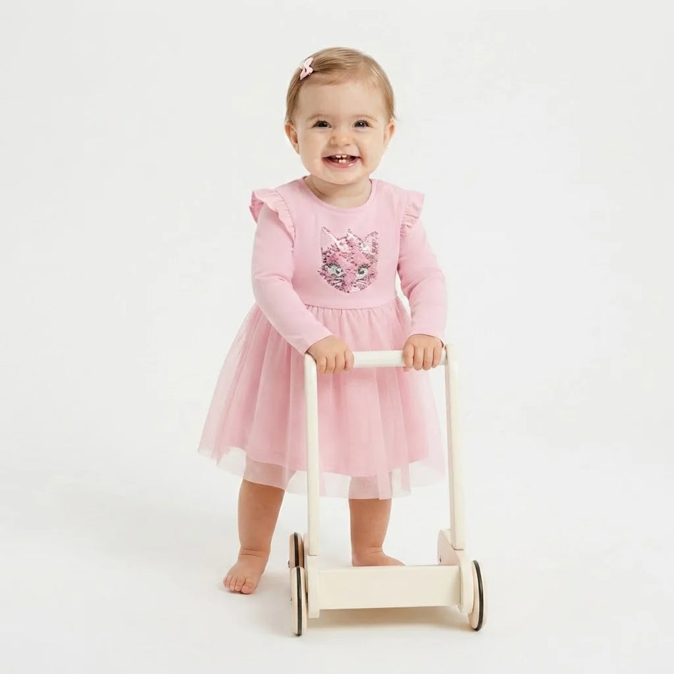 A smiling toddler stands barefoot on a white studio background, holding a small wooden push toy. The child wears a light pink long-sleeve dress with a tulle skirt and a sequined cat motif on the chest.