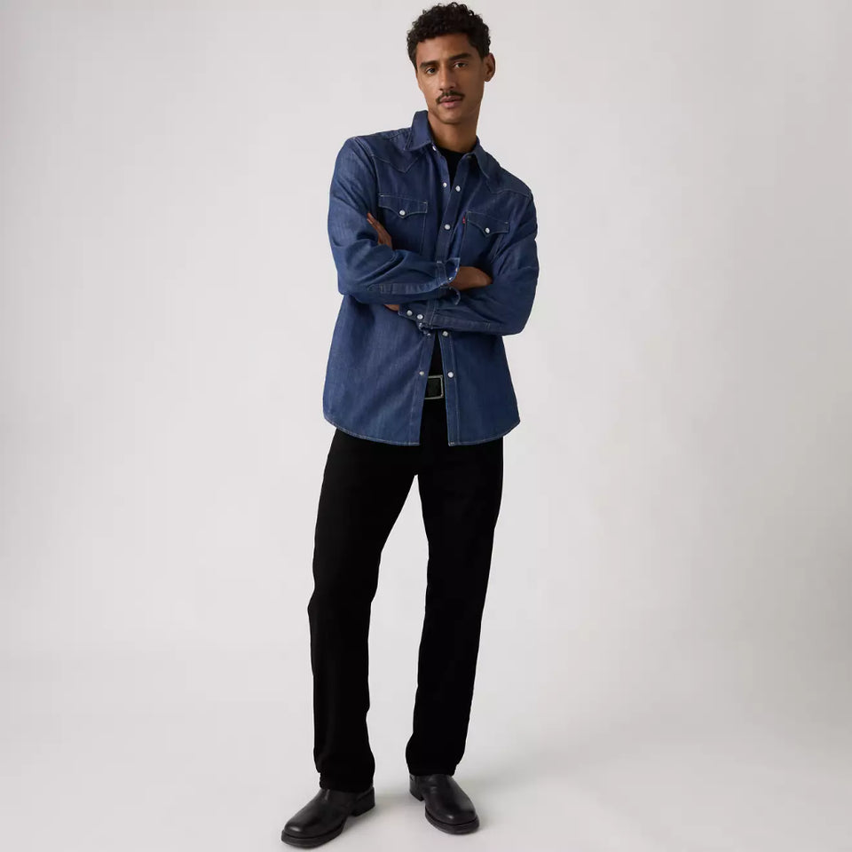 A man modeling a blue denim shirt with black pants and black leather shoes, standing with arms crossed against a light gray studio background.